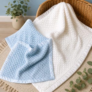 Könnte beinhalten: Weiche, strukturierte Babydecken in Weiß und Hellblau. Die Decken haben eine Satinborte und sind auf einem neutralfarbenen Teppich gefaltet. Ein Weidenkorb und eine kleine Topfpflanze befinden sich im Hintergrund.