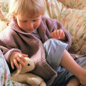 Può includere: Un bambino indossa un cardigan lavorato a maglia color malva con collo a scialle e chiusura con bottoni. Il bambino tiene in mano un giocattolo a forma di coniglietto di peluche. Lo sfondo presenta un tessuto a fantasia floreale.