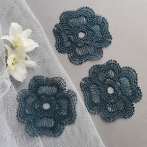 Op de afbeelding: Drie teal-kleurige, opengewerkte bloemvormige onderzetters. De onderzetters zijn gemaakt van een fijne, metallic draad en zijn gerangschikt op een grijze ondergrond. Witte orchideeën en een doorschijnende witte stof bevinden zich op de achtergrond.