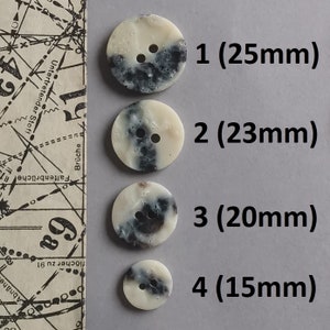 May include: Four white buttons with black speckles and two holes. The buttons are arranged in a row from largest to smallest. The buttons measure 25mm, 23mm, 20mm, and 15mm.