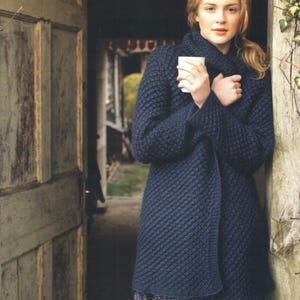 Peut inclure: Un cardigan bleu marine foncé à manches longues avec un motif en tricot diagonal texturé. Le vêtement possède un col montant et est porté enveloppé autour du torse. Une tasse en céramique blanche unie est tenue devant la poitrine.
