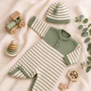 May include: A sage green and cream striped knitted romper with long sleeves, a buttoned placket, and a matching beanie featuring a small tassel. The set is accompanied by a wooden toy train, a stacking ring toy, a rattle, and a wooden star.