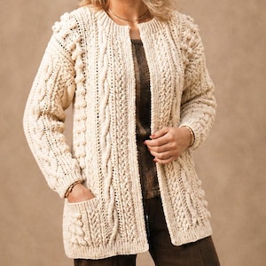 May include: Cream-colored, open-front cardigan sweater with a variety of cable knit and textured stitch patterns. The long-sleeved sweater features two front pockets and a round neckline. The garment is made of a thick, warm yarn.