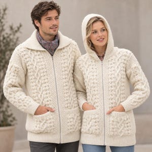 May include: Two cream-coloured zip-up cardigans made of thick knit wool. One cardigan features a wide collar, while the other includes an attached hood. Both garments display intricate cable knit patterns, textured bobble details, and two front pockets.
