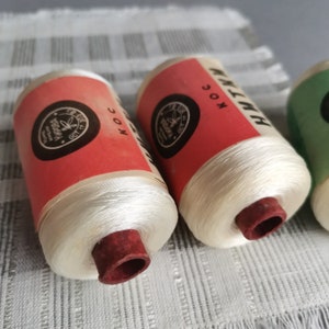 Puede incluir: Tres carretes de hilo blanco con etiquetas rojas y blancas. Las etiquetas tienen el texto "HOC" y "HMYK". Los carretes están sobre una superficie con un patrón blanco y gris.