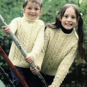 May include: Two children wearing cream-colored cable-knit sweaters. One sweater is a turtleneck, and the other is a button-down cardigan. The sweaters have a textured, woven design. The children are smiling and holding onto a metal bar.