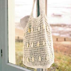 May include: Cream-colored, crocheted tote bag with a fish-shaped design. The bag is hanging by its handle in front of a window. The bag has a textured, open weave and is decorated with small, pale yellow fish appliqués.