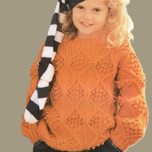 Puede incluir: Un suéter naranja tejido con un patrón de diamantes y bolitas texturizadas. El suéter tiene mangas largas y cuello redondo. También se ve un gorro a rayas blanco y negro.