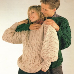May include: A beige cable-knit sweater with long sleeves and a crew neckline, worn alongside a dark green cable-knit sweater featuring long sleeves and a crew neckline. Both garments are constructed with a thick, textured yarn pattern.
