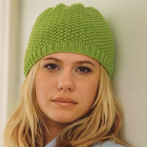 May include: A knitted green beanie hat with a ribbed brim. The hat is a bright, solid green colour and has a textured appearance. The hat is worn by a person with blonde hair.