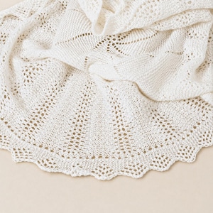 May include: A cream-colored knitted shawl featuring an intricate openwork lace pattern. The fabric displays a delicate, repeating geometric design with a scalloped edge that follows the curved hemline of the textile, showcasing fine yarn work.