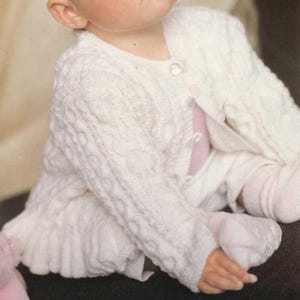 May include: A white long-sleeved knit cardigan featuring a textured cable knit pattern, a round neckline, and a front button closure. The garment is worn over a light pink shirt and is paired with matching white knit socks on the feet.