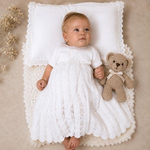 Könnte beinhalten: Ein weißes Strickkleid mit kurzen Ärmeln und einem zarten Spitzenmuster, getragen von einem Baby, das auf einem weißen Kissen und einer strukturierten weißen Decke liegt. Ein kleiner, hellbrauner gehäkelter Teddybär mit cremefarbenem Band liegt daneben.