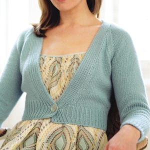 Peut inclure: Un gilet en tricot bleu clair doté d'une fermeture à bouton unique à la taille, de manches longues et d'une texture côtelée le long de l'ouverture avant et de l'ourlet. Le vêtement est porté sur une robe sans manches à motif jaune, vert et blanc.