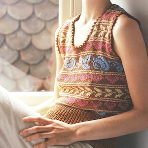 Op de afbeelding: Een mouwloze gebreide top in bruin, roze en blauw. De top heeft een V-hals en een patroon van bloemen en geometrische ontwerpen. Het kledingstuk is gemaakt van een zacht, getextureerd garen en lijkt handgemaakt.