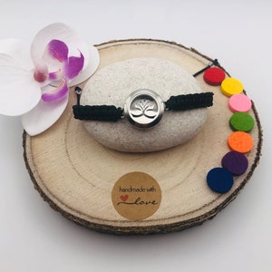 Puede incluir: Una pulsera de aromaterapia negra con un colgante plateado con un diseño de flor de loto. La pulsera se exhibe sobre una piedra gris clara, con almohadillas de fieltro de colores y una flor de orquídea blanca sobre una rodaja de madera. Una pequeña pegatina redonda dice "handmade with love".