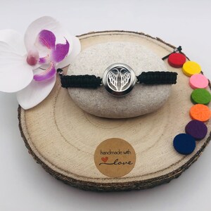 Puede incluir: Un brazalete de cordón negro con un dije plateado con alas de ángel. El brazalete se muestra sobre una superficie de madera con una pequeña etiqueta que dice "hecho a mano con amor".