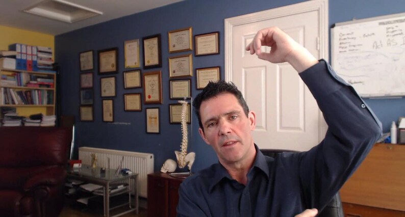 Peut inclure: Un homme portant une chemise bleue se tient dans un bureau avec un mur bleu. Il l&egrave;ve son bras droit au-dessus de sa t&ecirc;te. Il y a des certificats encadr&eacute;s sur le mur derri&egrave;re lui.