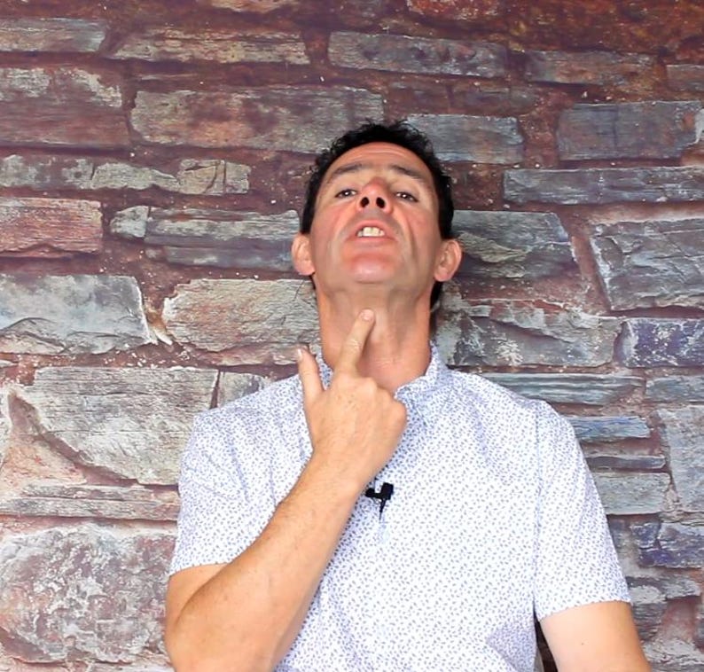May include: A man in a white button-down shirt with a small blue floral pattern points to his neck. The background is a stone brick wall.