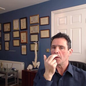 Peut inclure: Un homme aux cheveux courts bruns d&eacute;montre une technique avec son doigt sur son nez. Il porte une chemise bleue &agrave; col. L'arri&egrave;re-plan montre une pi&egrave;ce avec une biblioth&egrave;que, un bureau et un tableau blanc.