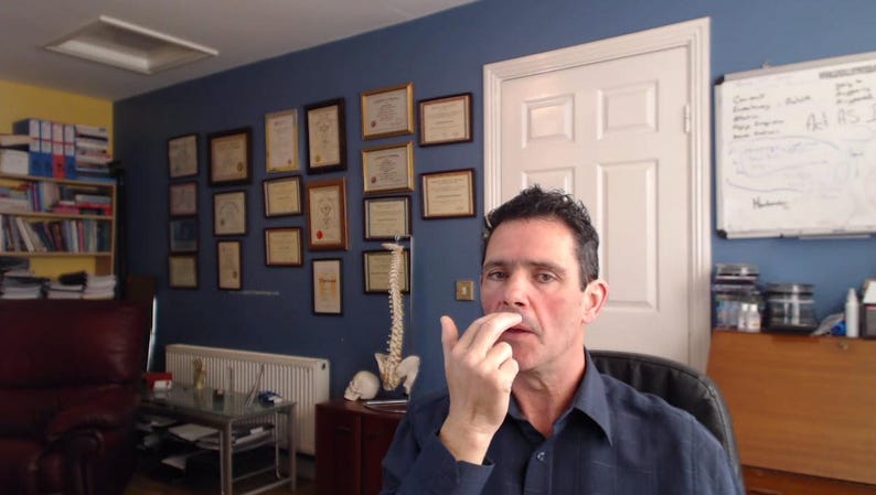 Peut inclure: Un homme aux cheveux courts bruns d&eacute;montre une technique avec son doigt sur son nez. Il porte une chemise bleue &agrave; col. L'arri&egrave;re-plan montre une pi&egrave;ce avec une biblioth&egrave;que, un bureau et un tableau blanc.