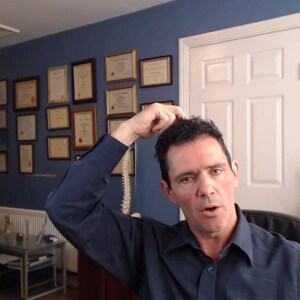 Peut inclure: Un homme aux cheveux fonc&eacute;s porte une chemise bleue et se tient dans un bureau. Il regarde la cam&eacute;ra et a la main sur la t&ecirc;te. Il y a des certificats encadr&eacute;s au mur derri&egrave;re lui.