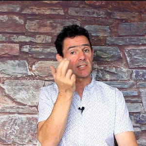 May include: A man wearing a white button-down shirt with a small blue pattern. He is touching his face with his fingers. The background is a brick wall.