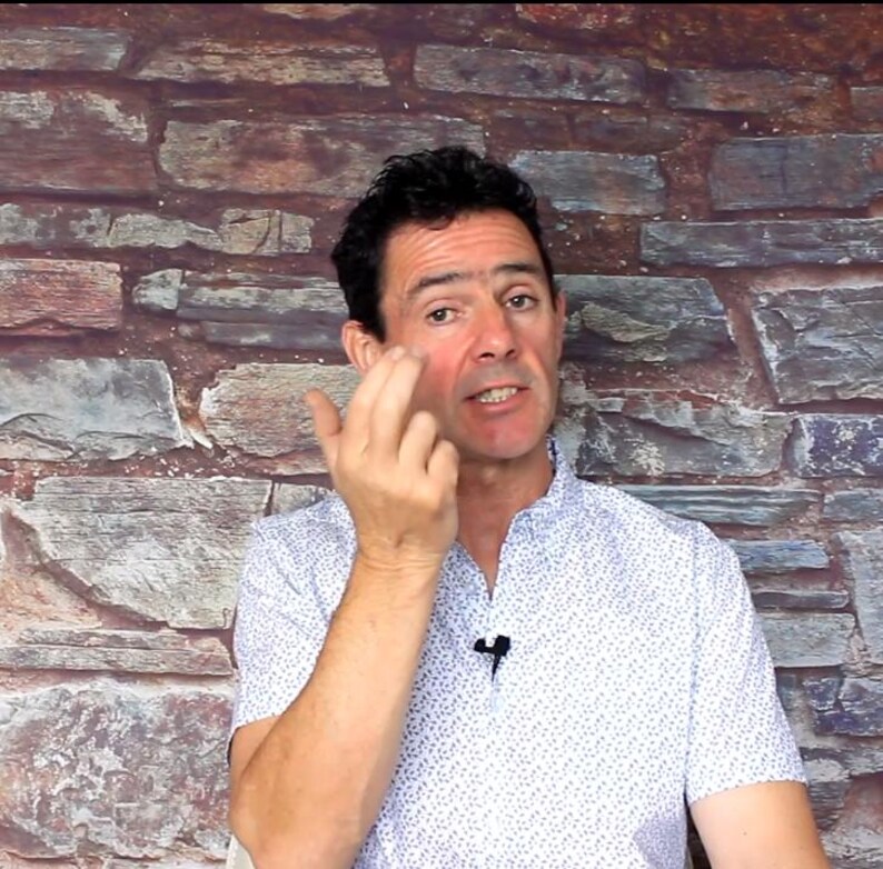 May include: A man wearing a white button-down shirt with a small blue pattern. He is touching his face with his fingers. The background is a brick wall.