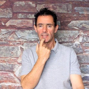 May include: A man wearing a gray polo shirt, posing in front of a brick wall. The man has dark hair and is touching his chin with his index finger, appearing thoughtful. The background features a mix of red, brown, and gray bricks.
