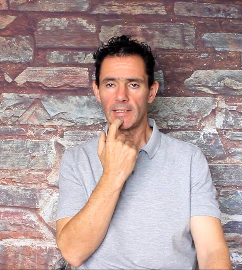 May include: A man wearing a gray polo shirt, posing in front of a brick wall. The man has dark hair and is touching his chin with his index finger, appearing thoughtful. The background features a mix of red, brown, and gray bricks.