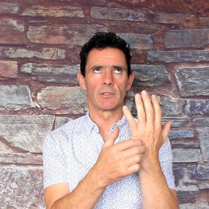 May include: A man wearing a white button-down shirt with a small blue pattern, looking upwards with a concerned expression. His right hand is raised, and he is touching his left wrist. The background is a brick wall.