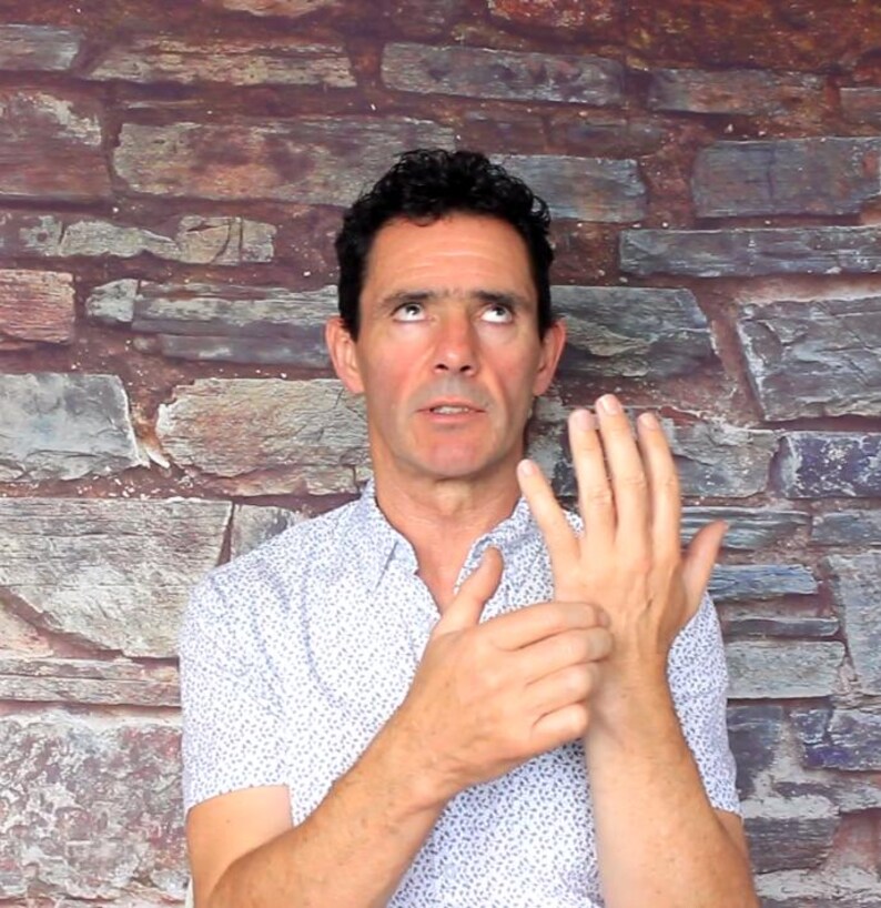 May include: A man wearing a white button-down shirt with a small blue pattern, looking upwards with a concerned expression. His right hand is raised, and he is touching his left wrist. The background is a brick wall.