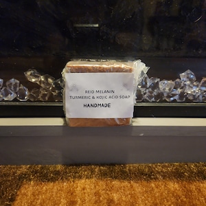 Può includere: Un sapone marrone avvolto in plastica trasparente con un'etichetta bianca che recita "Reid Melanin Turmeric & Kojic Acid Soap Handmade".