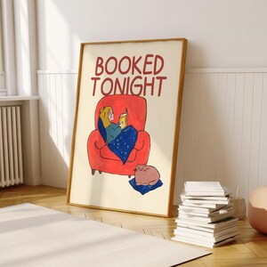 Puede incluir: Una impresi&oacute;n art&iacute;stica enmarcada con las palabras "BOOKED TONIGHT" en rojo. La ilustraci&oacute;n representa a una persona leyendo en un sill&oacute;n rojo, cubierta con una manta azul, con un gato cerca. La impresi&oacute;n est&aacute; en una habitaci&oacute;n con una alfombra blanca y una pila de libros.