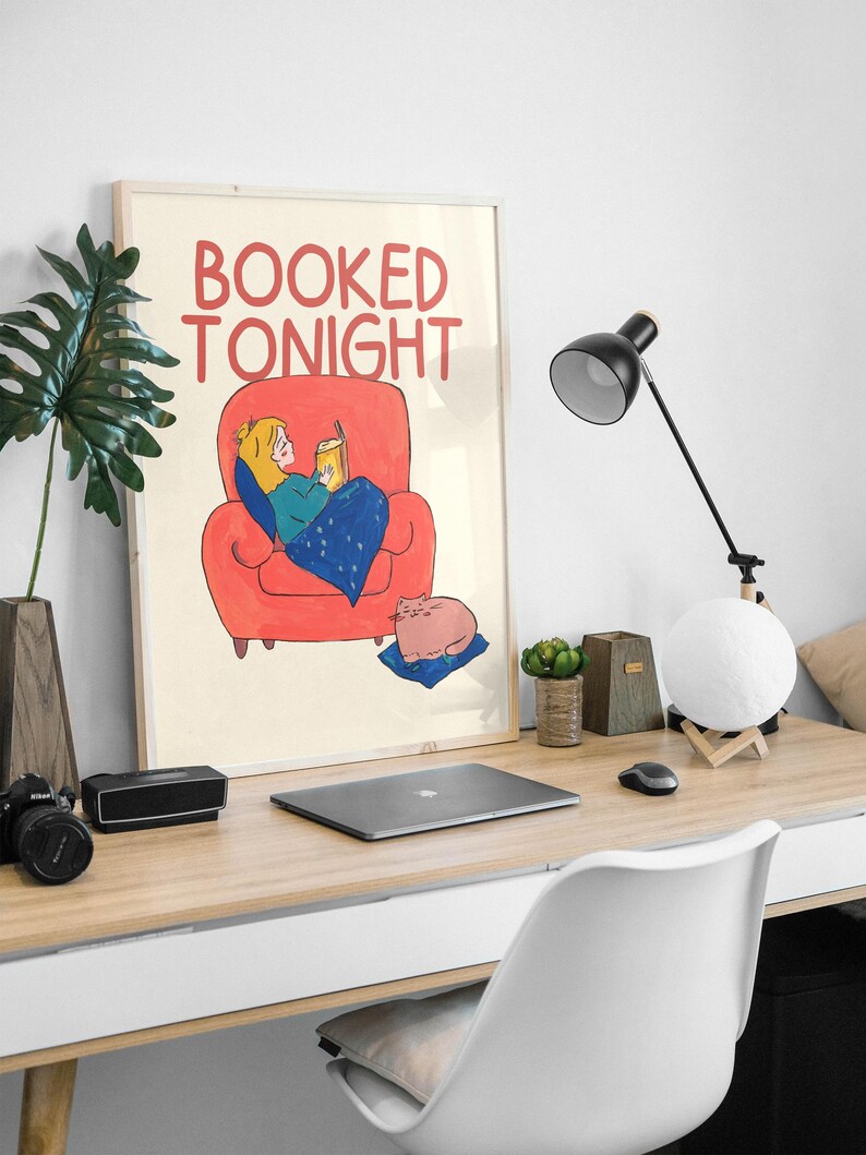 Puede incluir: Ilustraci&oacute;n enmarcada con el texto "BOOKED TONIGHT" que representa a una persona leyendo en un sill&oacute;n rojo, sosteniendo una bebida. Un gato rosa descansa sobre un coj&iacute;n azul. La escena se desarrolla en un escritorio de madera con un port&aacute;til, una l&aacute;mpara y accesorios.