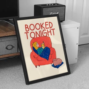Puede incluir: L&aacute;mina art&iacute;stica enmarcada con el texto "BOOKED TONIGHT" en rojo. La ilustraci&oacute;n representa a una persona leyendo en un sill&oacute;n rojo, cubierta con una manta azul, y un gato descansando sobre un coj&iacute;n azul.