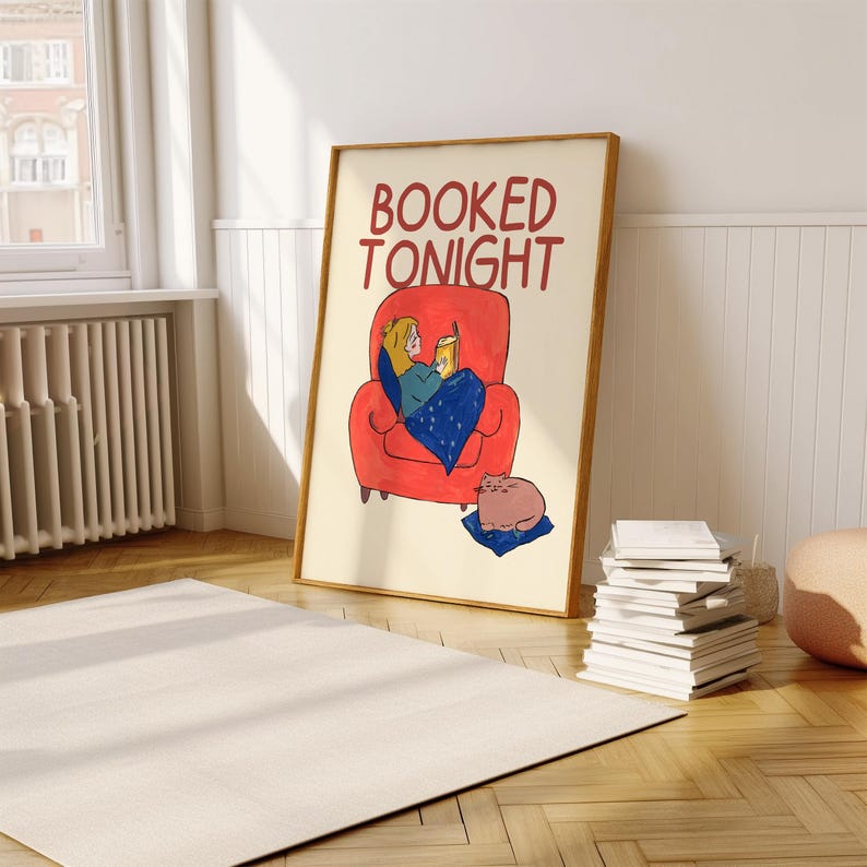 Puede incluir: Una impresi&oacute;n art&iacute;stica enmarcada con las palabras "BOOKED TONIGHT" en rojo. La ilustraci&oacute;n representa a una persona leyendo en un sill&oacute;n rojo, cubierta con una manta azul, con un gato cerca. La impresi&oacute;n est&aacute; en una habitaci&oacute;n con una alfombra blanca y una pila de libros.
