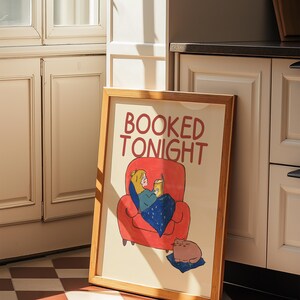 Puede incluir: Una impresi&oacute;n art&iacute;stica enmarcada con las palabras "BOOKED TONIGHT" en rojo. La ilustraci&oacute;n representa a una persona leyendo en un sill&oacute;n rojo, cubierta con una manta azul, y un gato descansando sobre un coj&iacute;n azul.