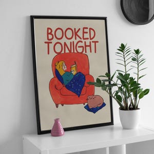 Puede incluir: Impresi&oacute;n art&iacute;stica enmarcada con la frase "BOOKED TONIGHT". La ilustraci&oacute;n muestra a una persona leyendo en un sill&oacute;n rojo, con un gato. Un jarr&oacute;n rosa y una planta en maceta est&aacute;n en una estanter&iacute;a blanca.