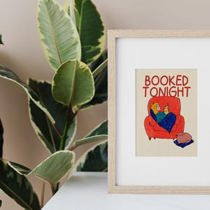 Puede incluir: L&aacute;mina enmarcada con el texto &laquo;BOOKED TONIGHT&raquo;. La ilustraci&oacute;n representa a una persona relaj&aacute;ndose en un sof&aacute; rojo, leyendo un libro bajo una manta azul. Un gato descansa sobre un coj&iacute;n azul. La impresi&oacute;n est&aacute; en un marco de madera clara.