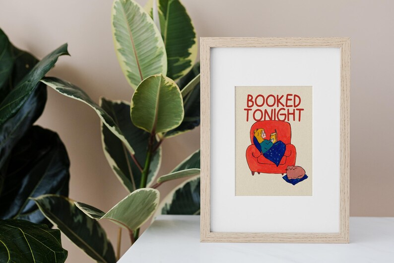 Puede incluir: L&aacute;mina enmarcada con el texto &laquo;BOOKED TONIGHT&raquo;. La ilustraci&oacute;n representa a una persona relaj&aacute;ndose en un sof&aacute; rojo, leyendo un libro bajo una manta azul. Un gato descansa sobre un coj&iacute;n azul. La impresi&oacute;n est&aacute; en un marco de madera clara.
