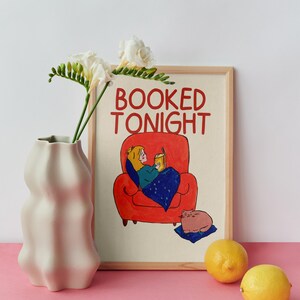 Puede incluir: Una impresi&oacute;n art&iacute;stica enmarcada con el texto "BOOKED TONIGHT" muestra a una persona leyendo en un sill&oacute;n rojo, un gato y una manta azul. Un jarr&oacute;n blanco con flores y dos limones est&aacute;n en primer plano. El fondo es blanco y la superficie es rosa.