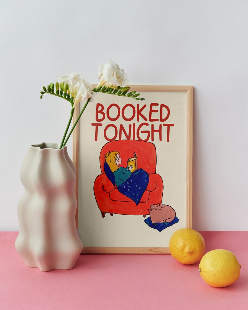 Puede incluir: Una impresi&oacute;n art&iacute;stica enmarcada con el texto "BOOKED TONIGHT" muestra a una persona leyendo en un sill&oacute;n rojo, un gato y una manta azul. Un jarr&oacute;n blanco con flores y dos limones est&aacute;n en primer plano. El fondo es blanco y la superficie es rosa.