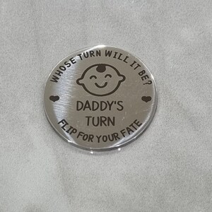 K&ouml;nnte beinhalten: Silberne M&uuml;nze mit dem Text "Whose turn will it be?" und "Daddy's turn" mit einem Cartoon-Babygesicht und zwei Herzen. Der Text "Flip for your fate" befindet sich unten auf der M&uuml;nze.