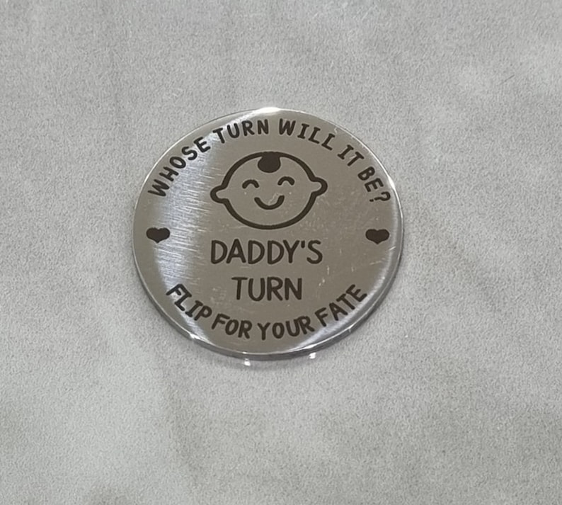 K&ouml;nnte beinhalten: Silberne M&uuml;nze mit dem Text "Whose turn will it be?" und "Daddy's turn" mit einem Cartoon-Babygesicht und zwei Herzen. Der Text "Flip for your fate" befindet sich unten auf der M&uuml;nze.