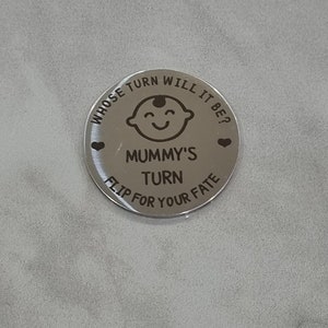K&ouml;nnte beinhalten: Eine silberne M&uuml;nze mit dem Text "Whose turn will it be?" und "Mummy's turn" mit einer Babygesichtsgrafik und Herzen. Der Text "Flip for your fate" befindet sich ebenfalls auf der M&uuml;nze.