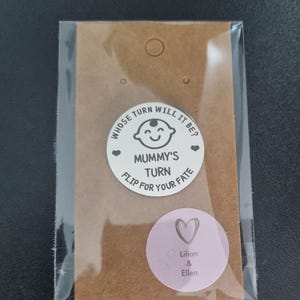 K&ouml;nnte beinhalten: Eine braune Papiert&uuml;te mit einem wei&szlig;en runden Aufkleber, auf dem "Whose turn will it be? Mummy's turn Flip for your fate" mit einem Cartoon-Babygesicht steht. Es gibt einen rosa runden Aufkleber mit einem Herzen und dem Text "Lilian & Ellen".