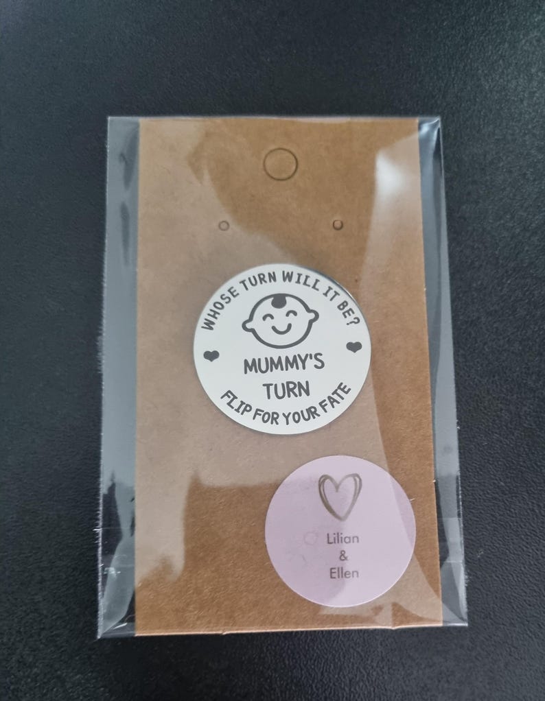 K&ouml;nnte beinhalten: Eine braune Papiert&uuml;te mit einem wei&szlig;en runden Aufkleber, auf dem "Whose turn will it be? Mummy's turn Flip for your fate" mit einem Cartoon-Babygesicht steht. Es gibt einen rosa runden Aufkleber mit einem Herzen und dem Text "Lilian & Ellen".