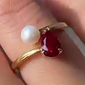 Op de afbeelding: Een gouden ring met een druppelvormige, dieprode edelsteen en een kleine, ronde witte parel. De ring heeft een delicaat, gedraaid bandontwerp.