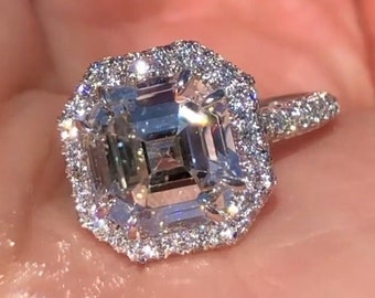 May include: A square-shaped diamond engagement ring with a halo setting. The ring is set in white gold and features a large, clear diamond in the center.